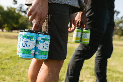 Person holding cans of 'THC Lite' brew in a park setting